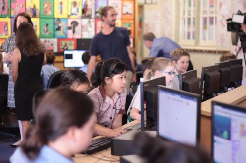 Photo of a Code Club in a classroom: six or seven children focus intently on Scratch programs and other tasks, and adults are helping and supervising in the background