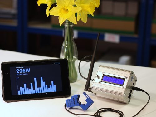 Photo: an emonPi energy monitoring unit in an aluminium case with an aerial and an LCD display, a mobile phone showing daily energy use as a histogram, and a bunch of daffodils in a glass bottle