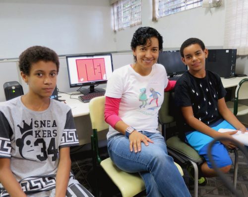 Two boys and a woman wearing a Code Club T-shirt sit and pose for the camera in a classroom