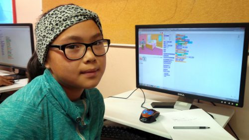 A girl sits by a desktop computer, with her Scratch coding project showing on the screen.