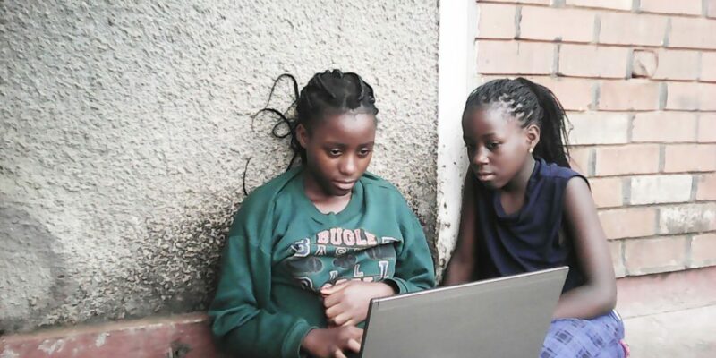 Two black girls sitting against an outside wall while working on a laptop