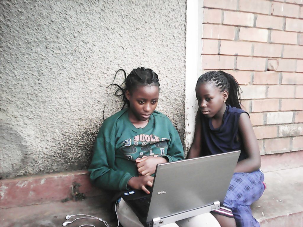 Two black girls sitting against an outside wall while working on a laptop