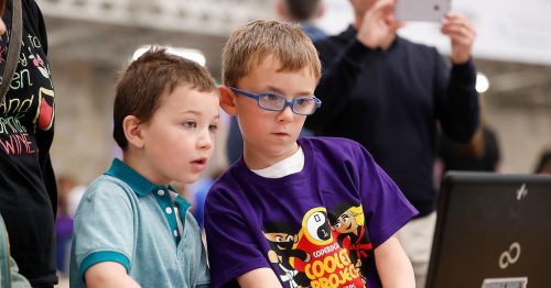 A young boy in a CoderDojo Ninja T-shirt shows another young boy his project, both concentrating intently