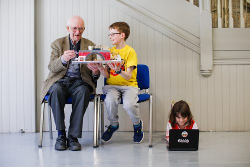  a boy showing a technology project to an old man, with a girl playing on a laptop on the floor — Coolest Projects North America