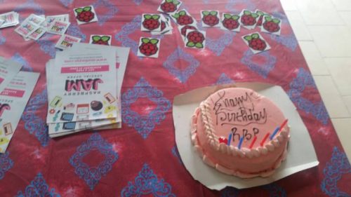 A round pink-iced cake decorated with the words "Happy Birthday RBP" and six candles, on a table beside Raspberry Pi stickers, Raspberry Jam stickers and Raspberry Jam fliers