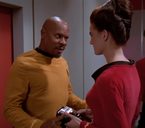 A still from an episode of Star Trek: Deep Space Nine: Jadzia Dax, holding an Original Series-sylte tricorder, speaks with Benjamin Sisko