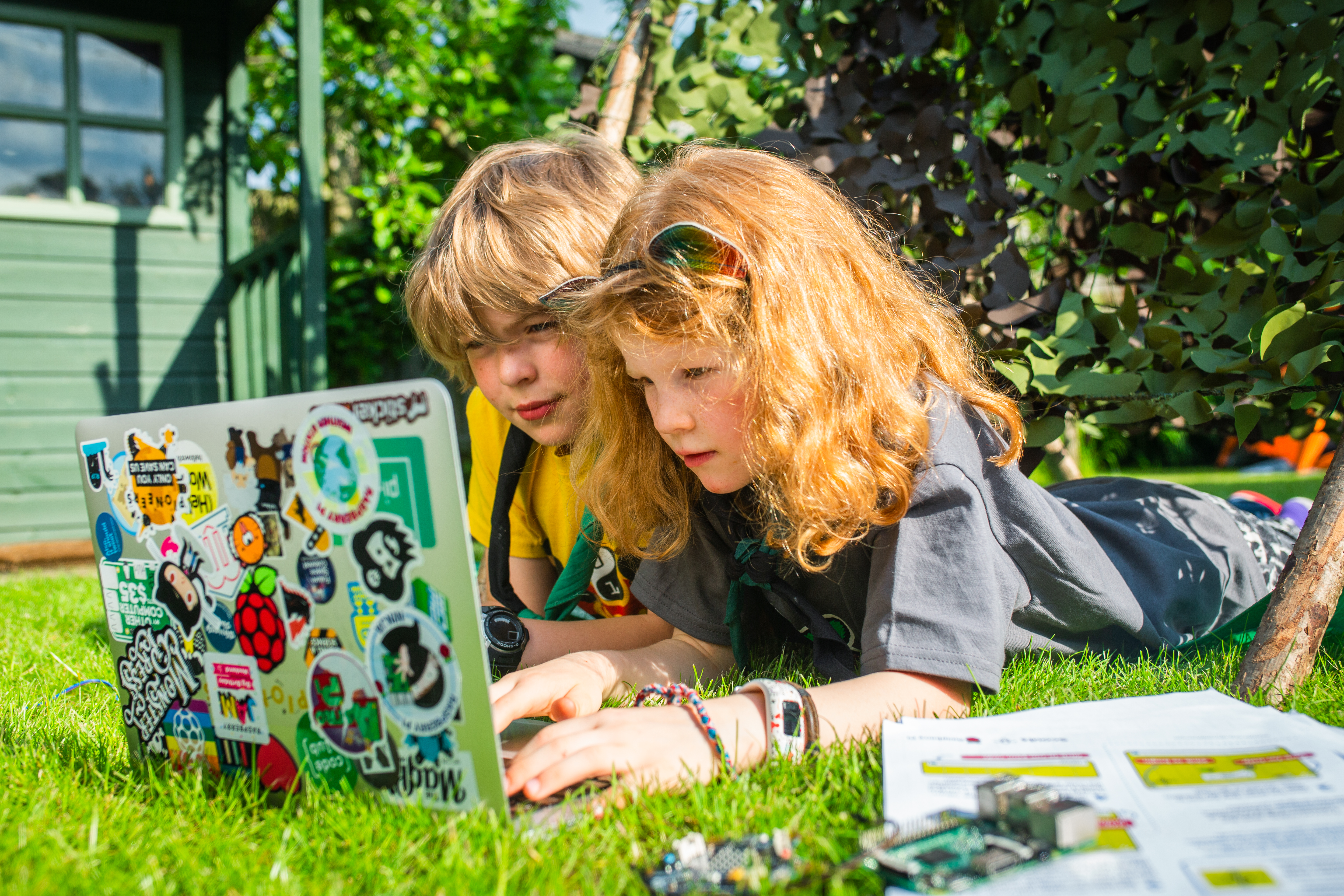 Two young children are propped on their elbows next to one another, using a laptop covered with stickers in a sunny garden. A Raspberry Pi and worksheets are beside them.