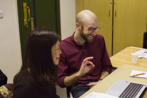 A woman and a man sit at a desk, evidently collaborating on work on a laptop. The woman is smiling and the man is grinning and making an "A-OK" hand gesture.