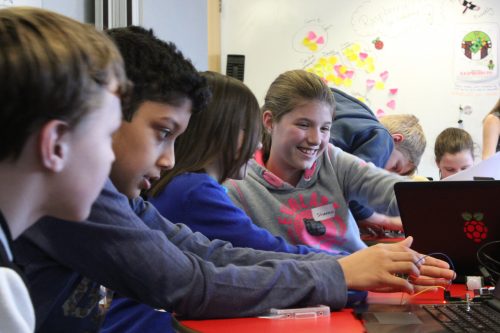 Several children, some smiling broadly and some concentrating intently, work with Raspberry PI computers and electronic components in a classroom