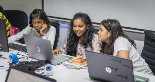 Three girls work at laptops. One is concentrating seriously. Two are smiling as they collaborate in their work.