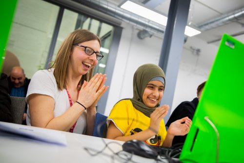 A young woman and a young girl sit side by side at a computer, grinning and clapping their hands in delight at what they are working on