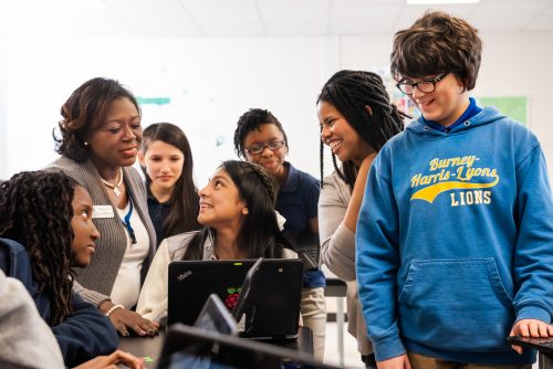 Two teachers and a group of learners are gathered around a laptop screen.