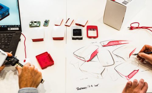 Top-down photo of a desk covered with white paper on which are a couple of Raspberry Pis and several cases. The hands of someone sketching red and white cases on the paper are visible. Also visible are the hands of someone measuring something with digital calipers, beside a laptop on the screen of which is a CAD model of a Raspberry Pi case.