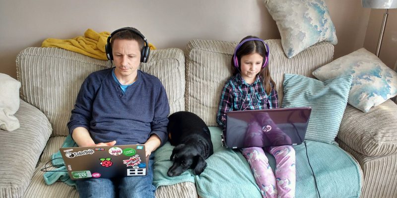 A girl and father sit on a sofa, each coding on their laptop, while a dog sleeps between them.