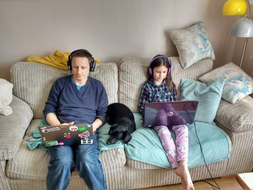 A girl has fun learning to code at home, sitting with a laptop on a sofa, with a dog sleeping next to her and her father writing code too.