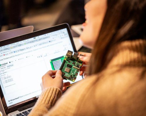 A woman holds up a Raspberry Pi computer in front of a laptop screen.