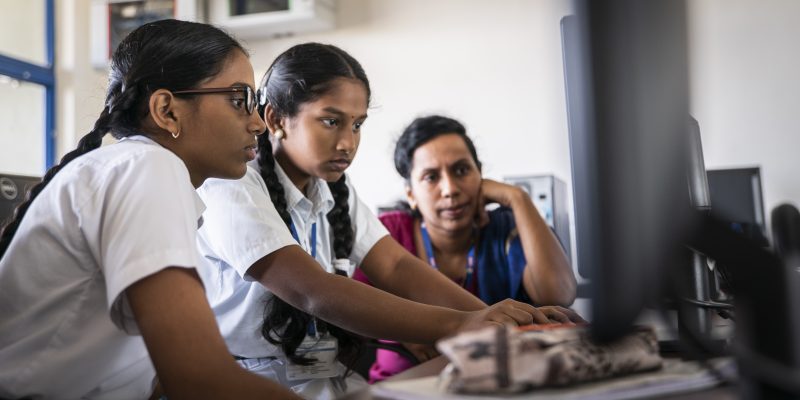 Two girls code at a desktop computer while a female mentor observes them.