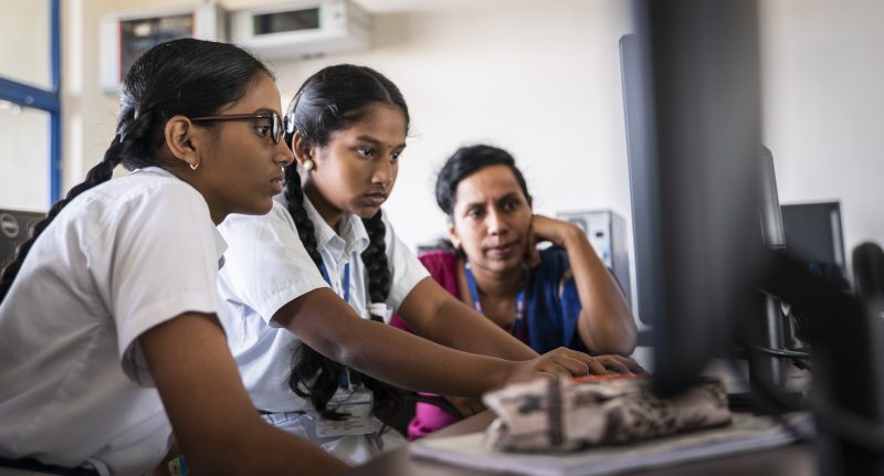 Two girls coding at a computer under supervision of a female teacher