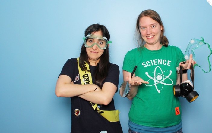 Dallas in a green t shirt stood next to Estefannie in a black t shirt on a blue background. Estefannie is wearing safety googles
