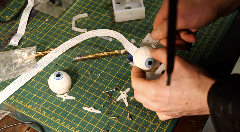 Maker inserting raspberry pi camera module inside a sliced ping pong ball. You can see the ribbons of the camera module sticking out of the ping pong ball half