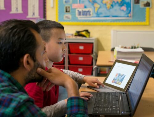 A teacher and a student work on a coding task at a laptop.