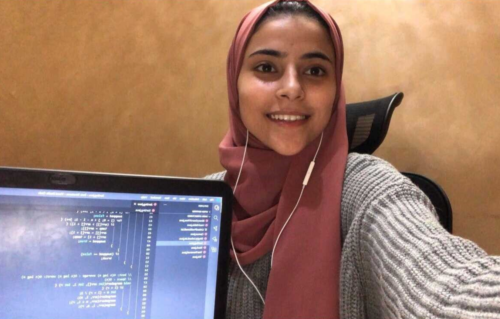 Young women in hijab smiles while holding up a laptop displaying code she has written