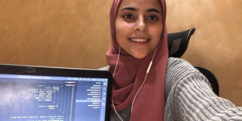 Young women in hijab smiles while holding up a laptop displaying code she has written