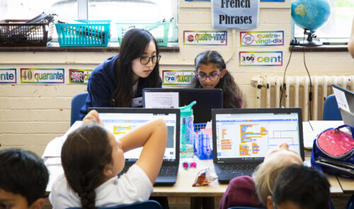 Computing classroom with woman teacher and young students at laptops doing Scratch coding.