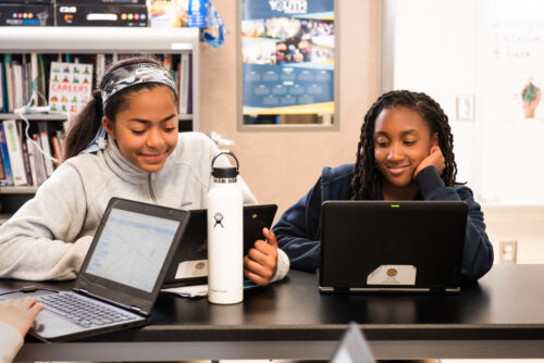 Two teenage girls do coding activities at their laptops in a classroom.