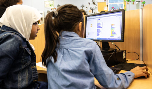 Two young girls, one of them with a hijab, do a Scratch coding activity together at a desktop computer.