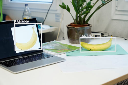 An image demonstrating that AI systems for object recognition do not distinguish between a real banana on a desk and the photo of a banana on a laptop screen.