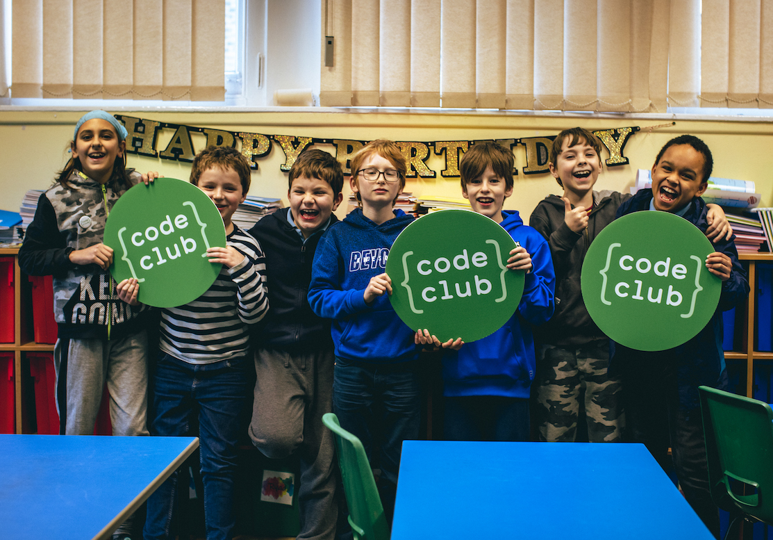 A group of smiling children hold up large cardboard Code Club logos.