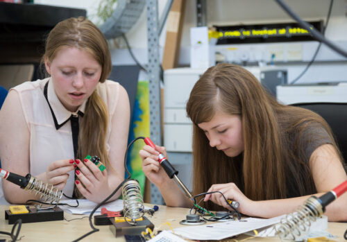 Two girls solder physical computing components in a workshop.