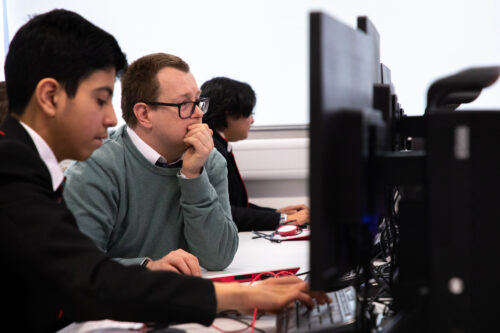 A computer science teacher sits with students at computers in a classroom.