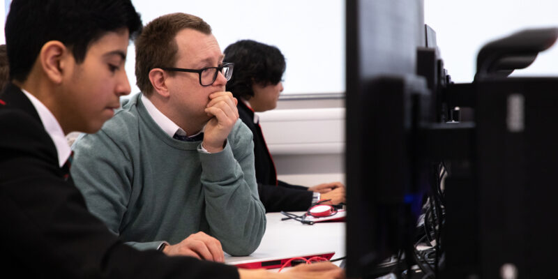 A computer science teacher sits with students at computers in a classroom.