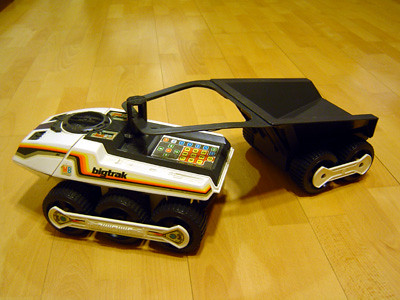 A Big Trak toy robot on wheels with a keypad on top and with a cart attached.