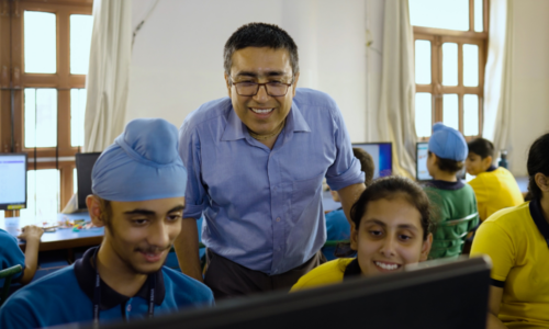 A group of students in a classroom being guided through their computing projects by a teacher.