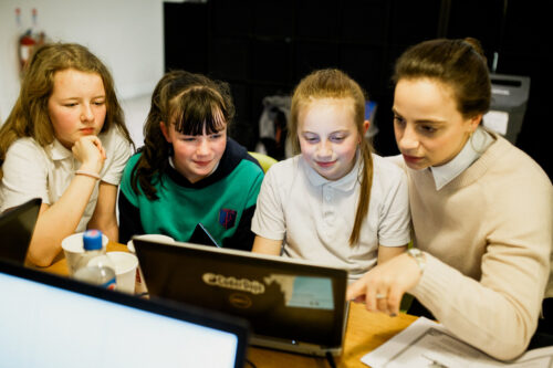 A teacher and three students in a classroom. The teacher is pointing at a computer screen. 