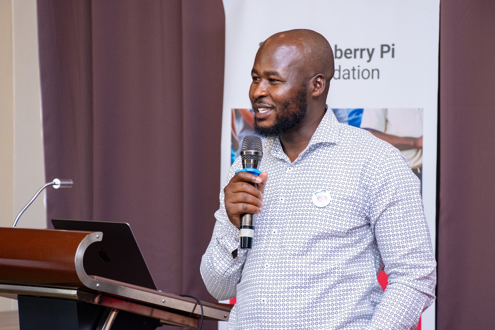 A speaker at the Raspberry Pi Foundation's Kenya Partner Showcase 2025, smiling and holding a microphone.