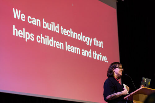 A person on stage stands before a slide with the sentence "We can build technology that helps children learn and thrive".