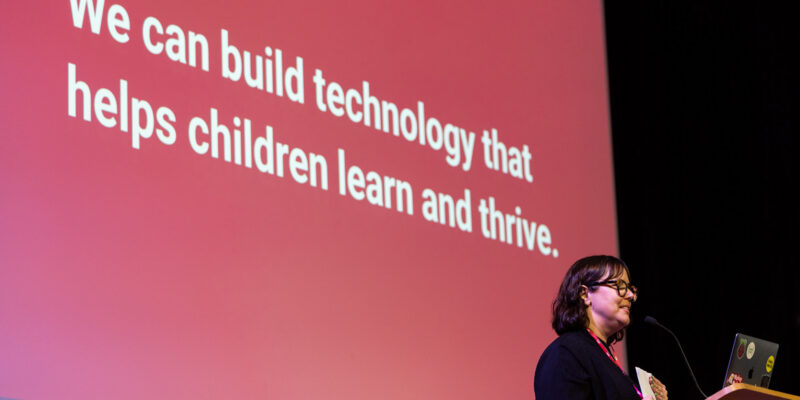 A person on stage stands before a slide with the sentence "We can build technology that helps children learn and thrive".