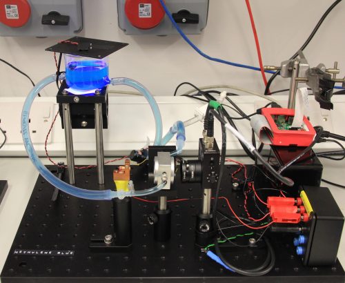 A laboratory photocatalysis setup at Cardiff University: on a bench are a beaker of water dosed with methylene blue "pollutant" under UV LED illumination, semi-transparent tubing connecting the contents of the beaker to a flow cell, a Raspberry Pi, and other components.