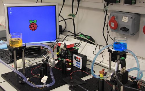 Laboratory photocatalysis setups at Cardiff University: on a bench are a large LCD display with a desktop showing the Raspberry Pi logo, beakers of water dosed with methyl orange and methylene blue "pollutants", semi-transparent tubing connecting the beakers' contents to flow cells, a Raspberry Pi, and other components.