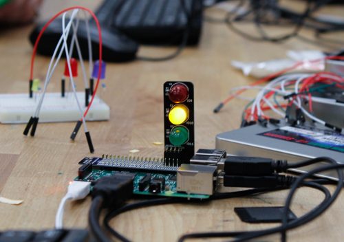 In the foreground, a Raspberry Pi computer with a small "traffic lights" board attached; it has red, yellow and green LEDs, and the yellow one is lit. In the background, various electronics components, a mouse and a keyboard.